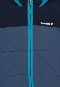 Doudoune bleu marine avec des accents teal contrastés, fermeture éclair complète et logo Timberland. Tissu texturé avec des sections de design matelassé.