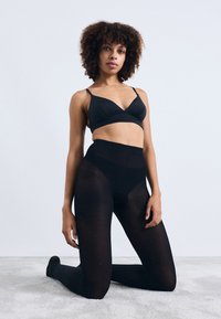 Frau mit lockigem Haar, die auf einem Teppich kniet und einen schwarzen BH sowie transparente schwarze Strumpfhosen mit kleinem Punktemuster trägt, vor einem weißen Hintergrund.