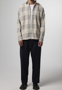Beige en grijs geruite flanellen shirt met knoopsluiting en twee voorzakken, gecombineerd met donkerblauwe, losse sweatpants en zwarte schoenen.