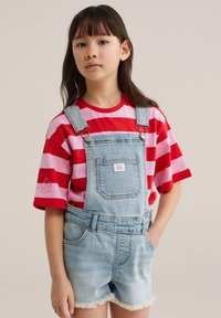 Noor tüdruk, kellel on pikk tume juuksed ja kes kannab punaseid ja roosaid triibulist särki, peal hele teksariidest lühikesed ülekuubikud, seisab ühe käega taskus.