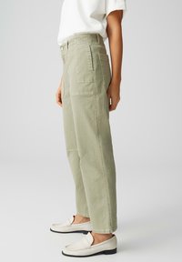 Vue de côté d'une personne portant un pantalon large vert clair avec poches appliquées, des mocassins blancs et un haut blanc à manches courtes rentré, sur un fond uni.