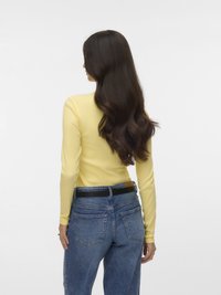 Kvinne med langt, bølgete mørkt hår iført en gul genser med lange ermer og blå jeans med svart belte, sett bakfra mot en hvit bakgrunn.