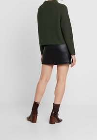 Frau trägt einen dunkelgrünen Strickpullover, einen schwarzen Leder-Minirock und braune, mit Absatz versehene Stiefeletten, steht vor einem schlichten Hintergrund.