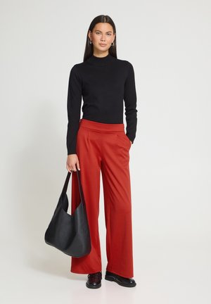 Femme portant un haut noir à manches longues et un pantalon rouge à jambes larges, tenant un grand sac à main noir, debout devant un fond clair uni.