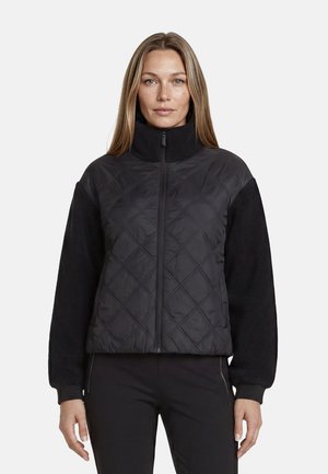 Femme portant une veste matelassée noire zippée avec des manches en polaire et un col montant, debout devant un fond clair uni.
