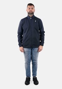 Giacca zip-up blu navy con collo alto, maniche lunghe e tasche laterali. Indossata sopra jeans azzurri chiari e sneakers nere.