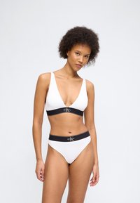 Γυναίκα με κατσαρά μαλλιά που φοράει λευκό μπούστο Calvin Klein και σλιπ με ψηλή γραμμή και μαύρες ελαστικές ταινίες με το λογότυπο, στέκεται μπροστά σε απλό φόντο.