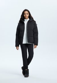 Schwarze Pufferjacke mit Kapuze, mit gesteppten Abschnitten, getragen über einem weißen Reißverschlussoberteil und schwarzen Hosen, kombiniert mit schwarzen Wanderschuhen.
