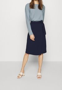 Námořnická zavinovací sukně se zavazováním na boku, zkombinovaná s světle modrým dlouhým rukávem. Modelka má na sobě lehké sandály na blokovém podpatku se tenkými pásky.