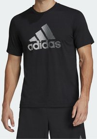 Lyhythihainen musta t-paita pehmeästä kankaasta, jossa on suuri vaakasuuntainen heijastava Adidas-logo edessä. Pyöreä kaula-aukko.