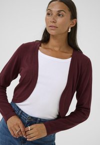 Cardigan court bourgogne en tissu tricoté doux, avec un design à ouverte sur le devant, des manches longues, porté avec un haut blanc et un jean bleu.