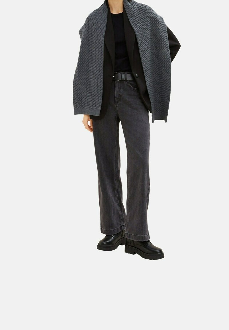 Personne portant un blazer noir, un top noir, un jean large gris foncé, une ceinture noire, des chaussures noires épaisses et une écharpe grise texturée drapée sur les épaules.