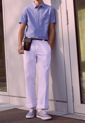 Homme portant une chemise bleu clair à manches courtes, un pantalon blanc avec des revers, des baskets grises et une pochette noire attachée à la ceinture, debout devant un bâtiment.