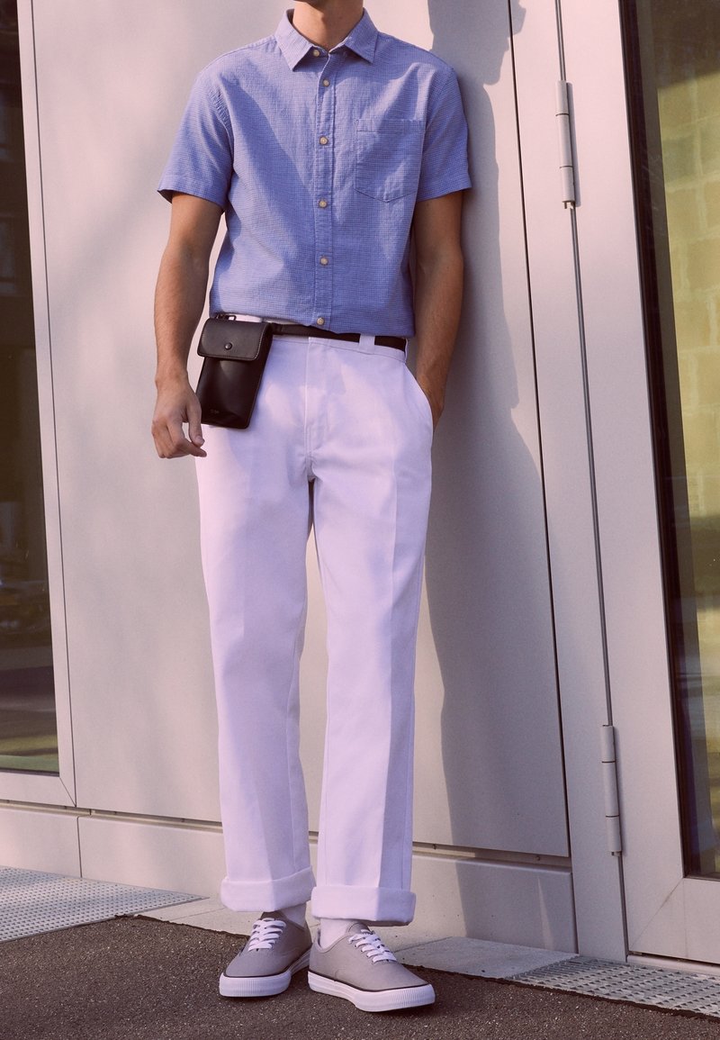 Homme portant une chemise bleu clair à manches courtes, un pantalon blanc avec des revers, des baskets grises et une pochette noire attachée à la ceinture, debout devant un bâtiment.