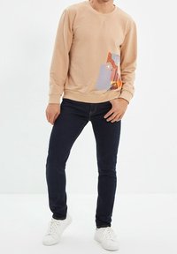Sudadera beige con cuello redondo, con patrones geométricos y texturas coloridas en el lado inferior izquierdo, combinada con jeans oscuros y zapatillas blancas.
