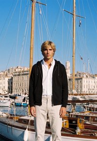 Jeune homme aux cheveux blonds portant une chemise blanche et une veste noire, debout devant des voiliers amarrés dans une marina sous un ciel bleu clair.