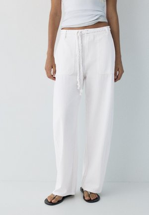 Osoba nosząca białe spodnie o szerokich nogawkach z przednimi kieszeniami i plecionym paskiem, w połączeniu z czarnymi klapkami na białej podłodze.