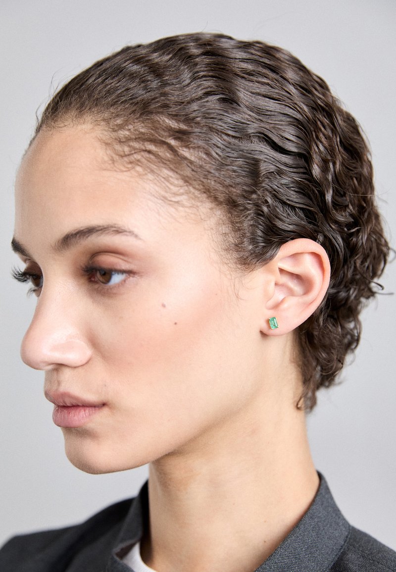 Σκουλαρίκια σε σχήμα ζαφειριού σε πράσινο χρώμα, τοποθετημένα σε μια απλή μεταλλική βάση, φορεμένα σε κοντινό προφίλ ενός ατόμου με κυματιστά μαλλιά και ουδέτερο μακιγιάζ.