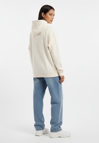 Felpa con cappuccio color crema dal taglio morbido, abbinata a jeans dritti in denim chiaro e sneakers bianche. Texture liscia, dettagli minimi.