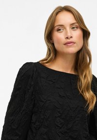 Top nero in tessuto con texture, caratterizzato da un collo a giro e maniche lunghe con dettagli arricciati. Il tessuto presenta un motivo stropicciato in tutta la sua superficie.