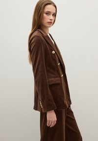 Bruine fluwelen double-breasted blazer met gouden knopen, zijzakken en een getailleerde pasvorm, getoond op een vrouw tegen een effen achtergrond.