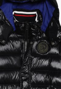 Veste noire rembourrée avec une finition brillante, intérieur bleu, fermeture éclair et un patch rond vert représentant un design de tête sur la poitrine.