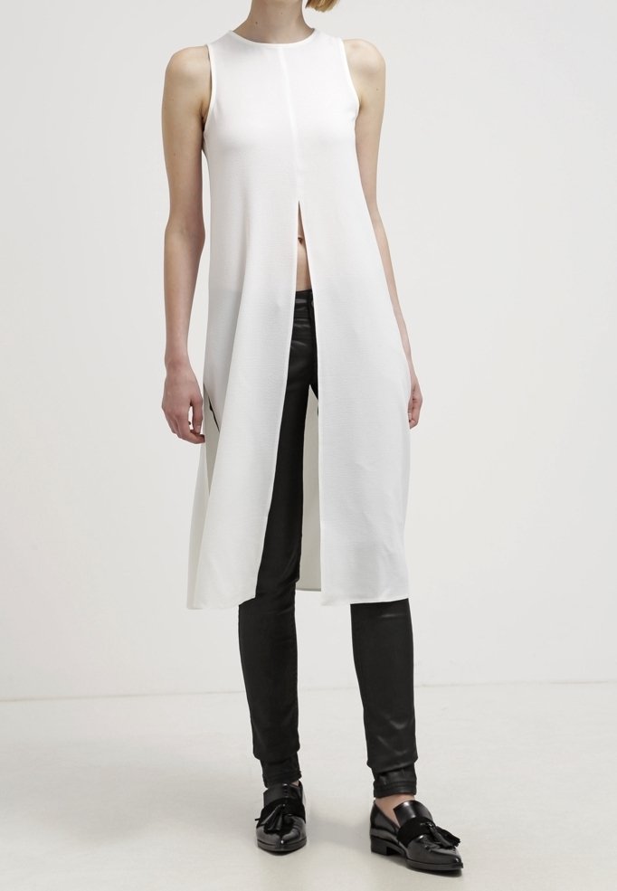 Robe blanche sans manches avec un design à fente avant, faite de tissu texturé. Assortie à un pantalon noir ajusté et des chaussures à enfiler.