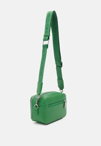 Zelená kožená kabelka přes rameno se stříbrnou hardwarou, obdélníkový tvar, zipové kapsy a nastavitelný popruh s názvem značky.