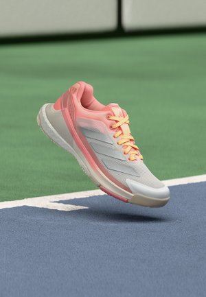 Zapatilla deportiva de color gris claro y rosa con cordones amarillos flotando sobre la línea de una pista de tenis en una superficie azul y verde.