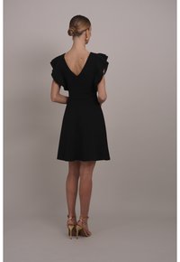 Schwarzes Kleid mit kurzen Rüschenärmeln, einem V-Ausschnitt hinten, tailliertem Oberteil und ausgestelltem Rock. Getragen mit goldenen, schlichten High Heels.