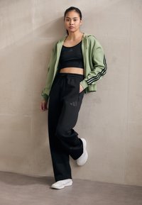 Žalias zip-up džemperis su juodomis juostelėmis virš juodos trumpomis rankovėmis bluzoną, derinamas su juodomis kelnėmis ir baltais sportiniais bateliais, prieš neutralią fono.