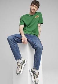 Camiseta verde de manga corta con un pequeño logo naranja, combinada con jeans azules y zapatillas grises y beige. Diseño simple; corte relajado.