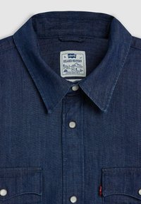 Camisa de mezclilla en azul oscuro con un cierre de botones en la parte delantera, cuello puntiagudo y dos bolsillos en el pecho. Cuenta con botones a presión plateados y una etiqueta con el logo.