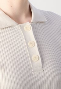Chemise beige clair à col avec une texture côtelée, dotée de trois boutons crème au niveau du col et de détails de couture subtils.