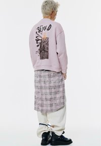 Felpa rosa chiaro con design grafico sulla schiena, sovrapposta a una gonna di plaid lunga fino al ginocchio. Pantaloni larghi color crema con stivali neri.