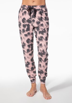 Frau trägt rosa-schwarze Joggerhose mit Leopardenmuster, elastischen Bündchen und schwarzem Kordelzug, steht barfuß auf weißem Boden.