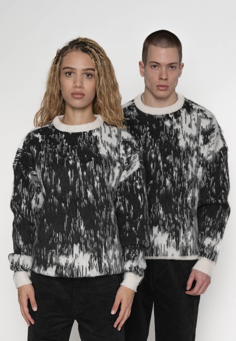 Dos modelos con suéteres de patrones abstractos en blanco y negro a juego y pantalones negros, posando frente a un fondo blanco liso.