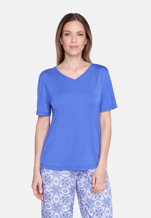 Frau mit glatten hellbraunen Haaren, die ein blaues T-Shirt mit V-Ausschnitt und kurzen Ärmeln sowie gemusterte blaue und weiße Hosen trägt und vor einem weißen Hintergrund steht.