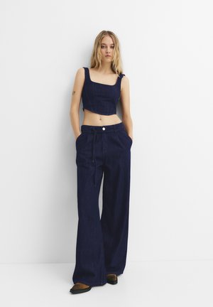 Top scurt din denim de culoare bleumarin, cu decolteu pătrat, asortat cu pantaloni largi bleumarin. Ambele piese au cusături vizibile și o croială lejeră.