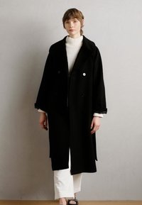 Manteau en laine noir avec un devant croisé et de grands revers, associé à un pull col roulé côtelé blanc et à un pantalon blanc. Texture lisse et soyeuse.