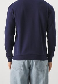 Felpa blu navy con maniche lunghe e orlo a coste, realizzata in un tessuto morbido, con un design semplice. Un paio di pantaloni azzurri chiari sullo sfondo.