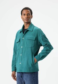 Groene fleecejas met een kraag, twee borstzakken en een sluiting met drukknopen. Zachte textuur, losse pasvorm en lange mouwen.