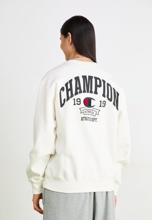 Bela športna majica s kapuco, na hrbtni strani je natisnjeno "CHAMPION" in "1919" v črni barvi. Ima rebraste manšete in rob, sproščen kroj ter okroglo izrez.