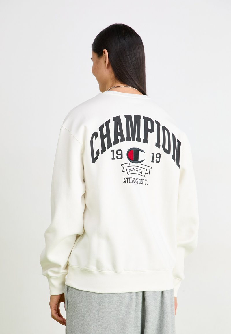Bijela dukserica s natpisom "CHAMPION" i "1919" ispisanim crnom bojom na leđima. Ima rebraste manžete i donji rub, s opuštenim krojem i okruglim izrezom.