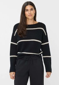 Zwarte gebreide trui met beige horizontale strepen, wijde mouwen en een ontspannen pasvorm, gecombineerd met zwarte broek met trekkoord.
