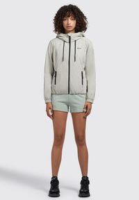 Lichtgrijze, met rits gesloten jas met een capuchon, zwarte accenten en zijzakken, gedragen over mintgroene shorts en zwarte schoenen. Gladde stoftextuur.