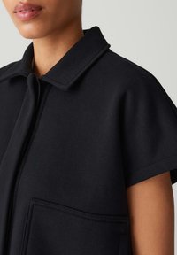 Schwarzes Baumwollmisch-T-Shirt mit kurzen Ärmeln, flachem Kragen, Knopfleiste vorne und einer einzelnen aufgesetzten Tasche, mit glatter Textur und komfortablem Schnitt.