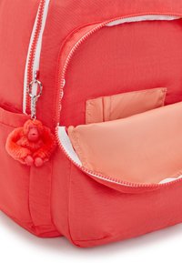 Mochila de nylon color coral con un bolsillo frontal con cremallera, acentos de cremallera blanca y un llavero de gorila rojo esponjoso adjunto.