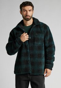 Veste chemise en tartan flou, teal et noir, avec un devant à boutons, un col et une poche poitrine. Elle présente un tissu texturé et une coupe chaleureuse et décontractée.