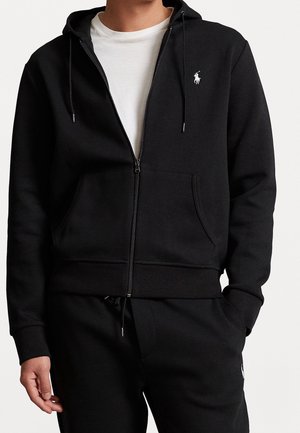 Man draagt een zwarte hoodie met ritssluiting en bijpassende zwarte joggingbroek met wit logo, witte t-shirt eronder, hand in zak.